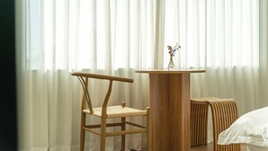 A room with a wooden chair, a small round table with a vase, and a bed with white bedding.