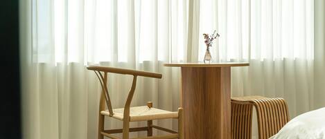 Una habitación con una silla de madera, una mesita redonda con un florero y una cama con ropa de cama blanca.