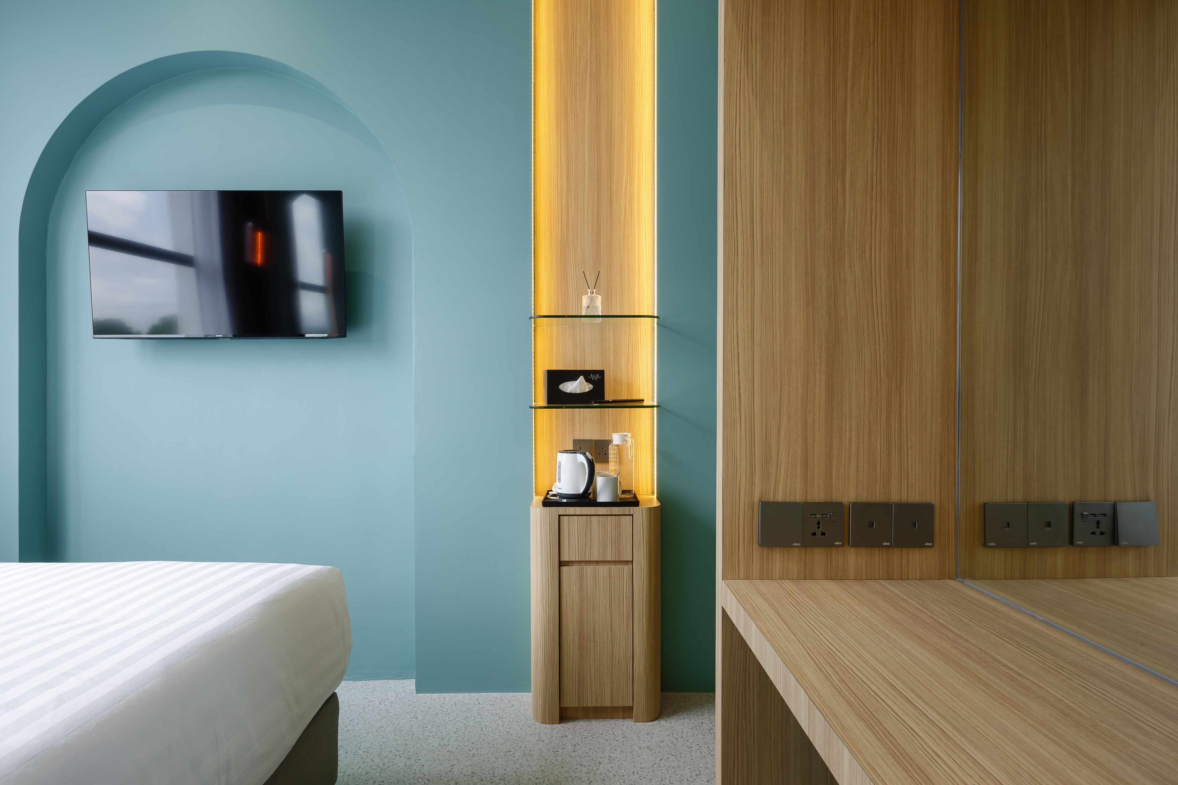 A modern hotel room with a bed, a TV mounted on the wall, a small wooden cabinet with a coffee maker, and a wooden wardrobe with built-in outlets.