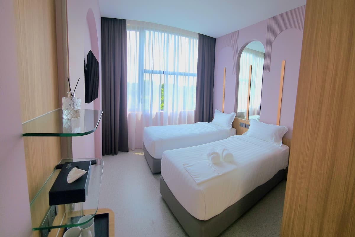 A hotel room with two beds, a glass desk with a cup and a remote control, and a large window with curtains.