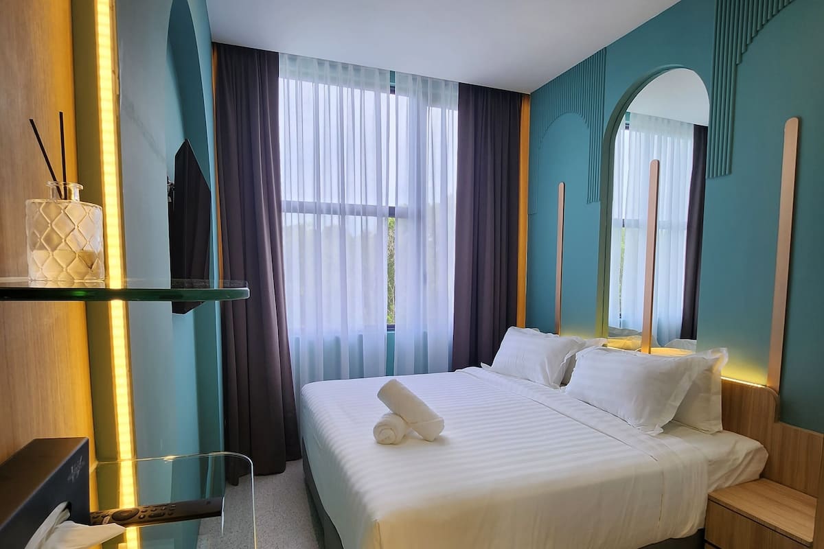 A modern hotel room with a neatly made bed, a glass side table with a remote control, and a view of the outdoors through sheer curtains.