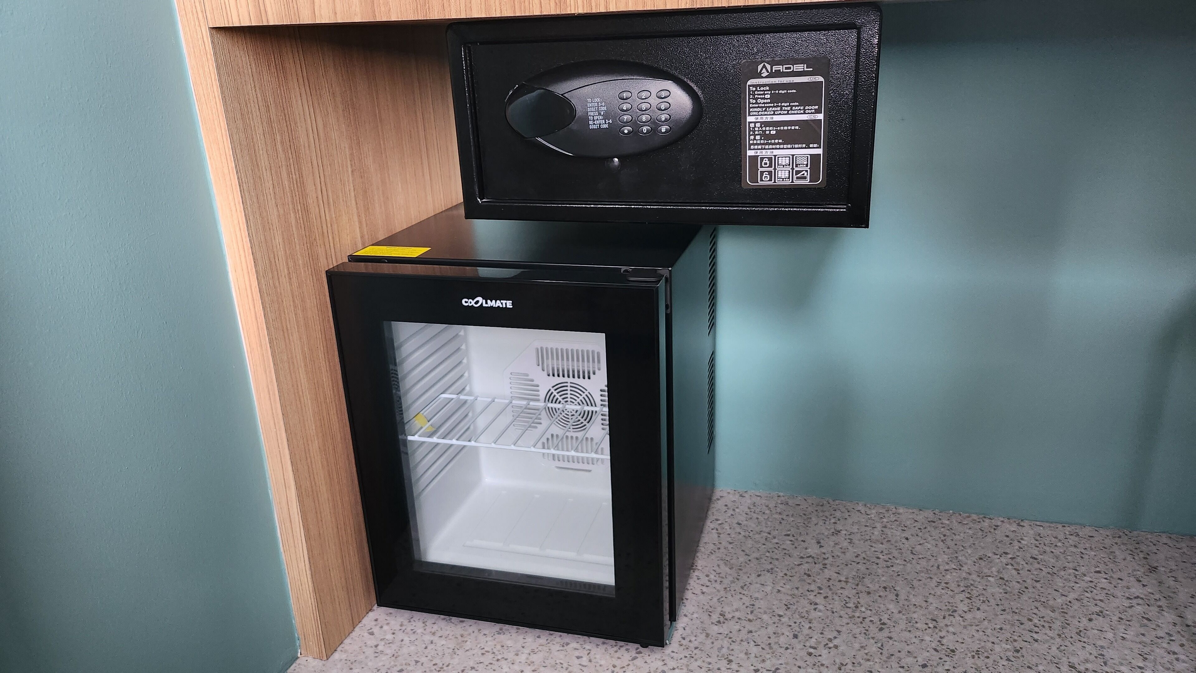 A compact refrigerator with a digital lock and a glass door, placed in a corner with a wooden cabinet above it.