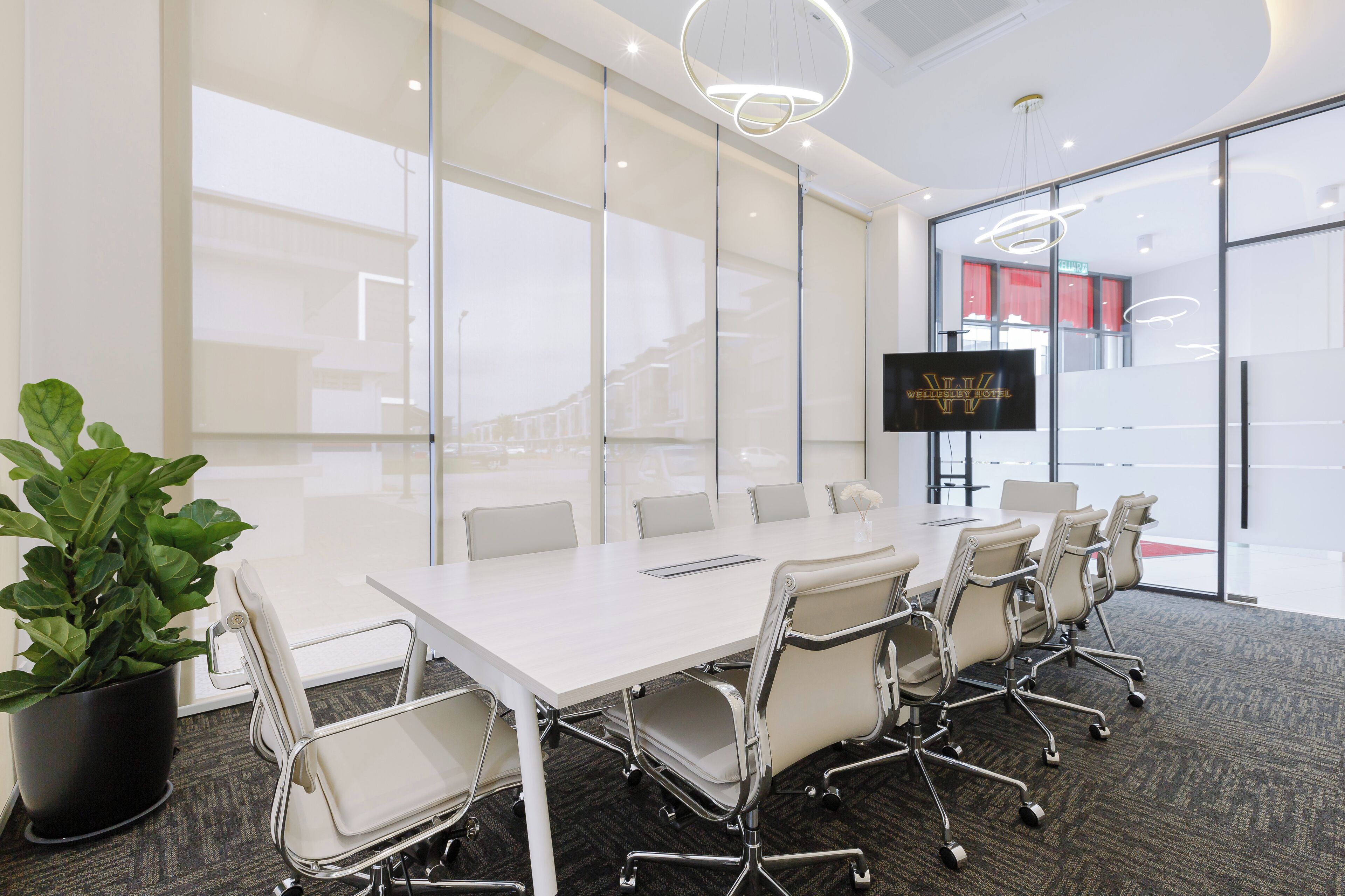 A conference room with a large table, white chairs, a potted plant, and a TV screen displaying a hotel name.