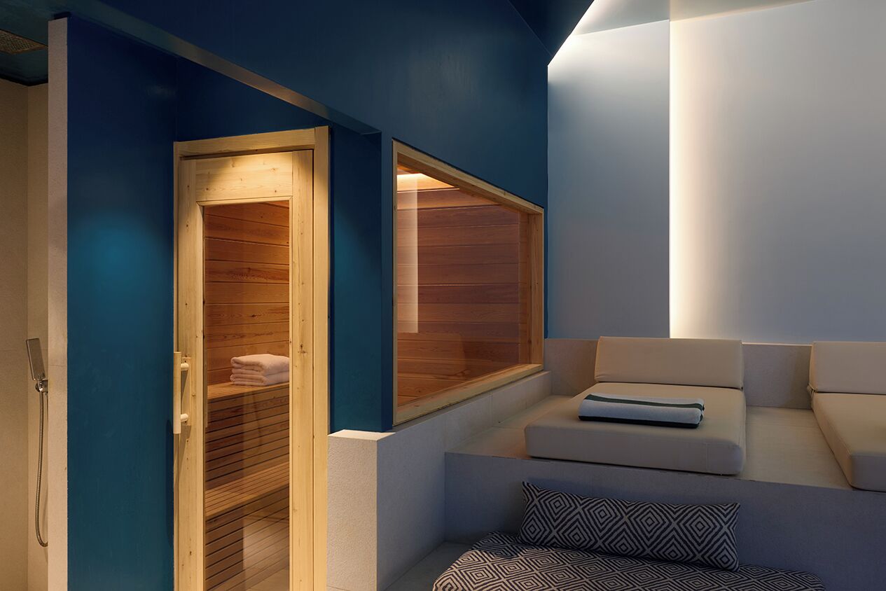 A modern spa room with a wooden sauna, a seating area with cushions, and a large window.