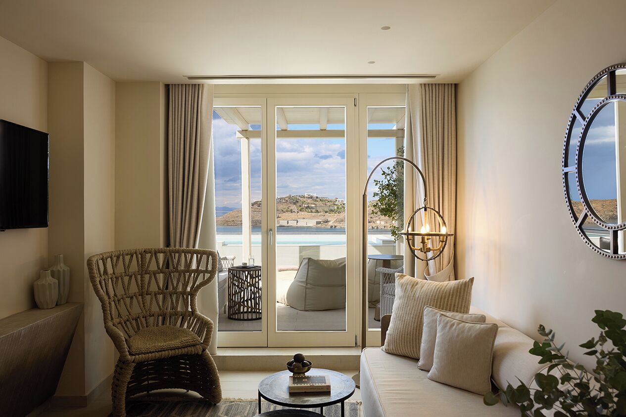 A room with a sofa, a wicker chair, a small table, a TV, and a view of the sea through large glass doors.