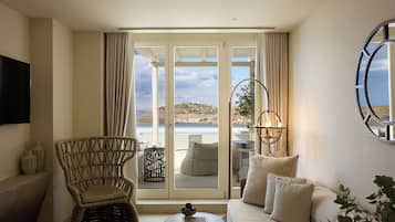 A room with a sofa, a wicker chair, a small table, a TV, and a view of the sea through large glass doors.