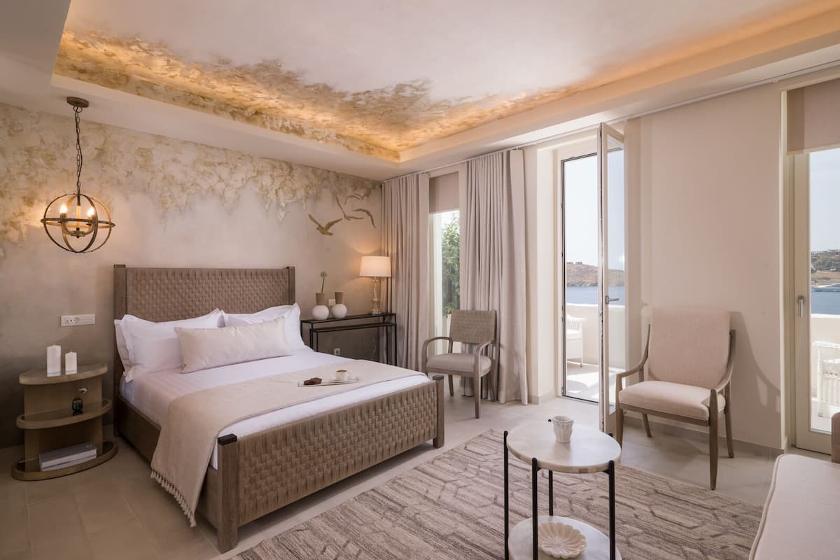 Een hotelkamer met een groot bed, een klein tafeltje, twee stoelen, een nachtkastje en uitzicht op zee door een schuifdeur.