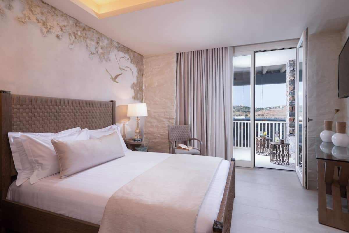 Een hotelkamer met een groot bed, een nachtkastje met lamp, een stoel, een balkon met buitenmeubilair en een televisie op een kast.