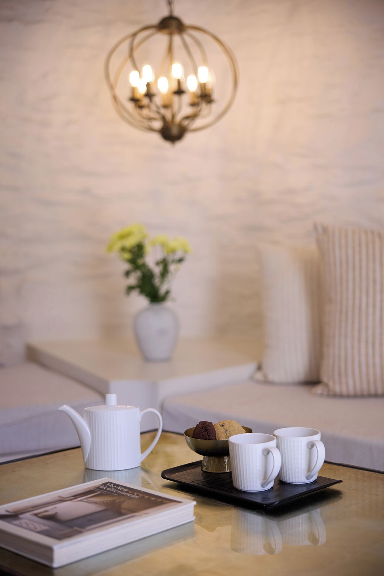 Un rincón acogedor con un sofá blanco, una mesita baja con una bandeja que contiene una tetera, dos tazas y galletas. Un jarrón con flores amarillas está en la pared detrás del sofá.