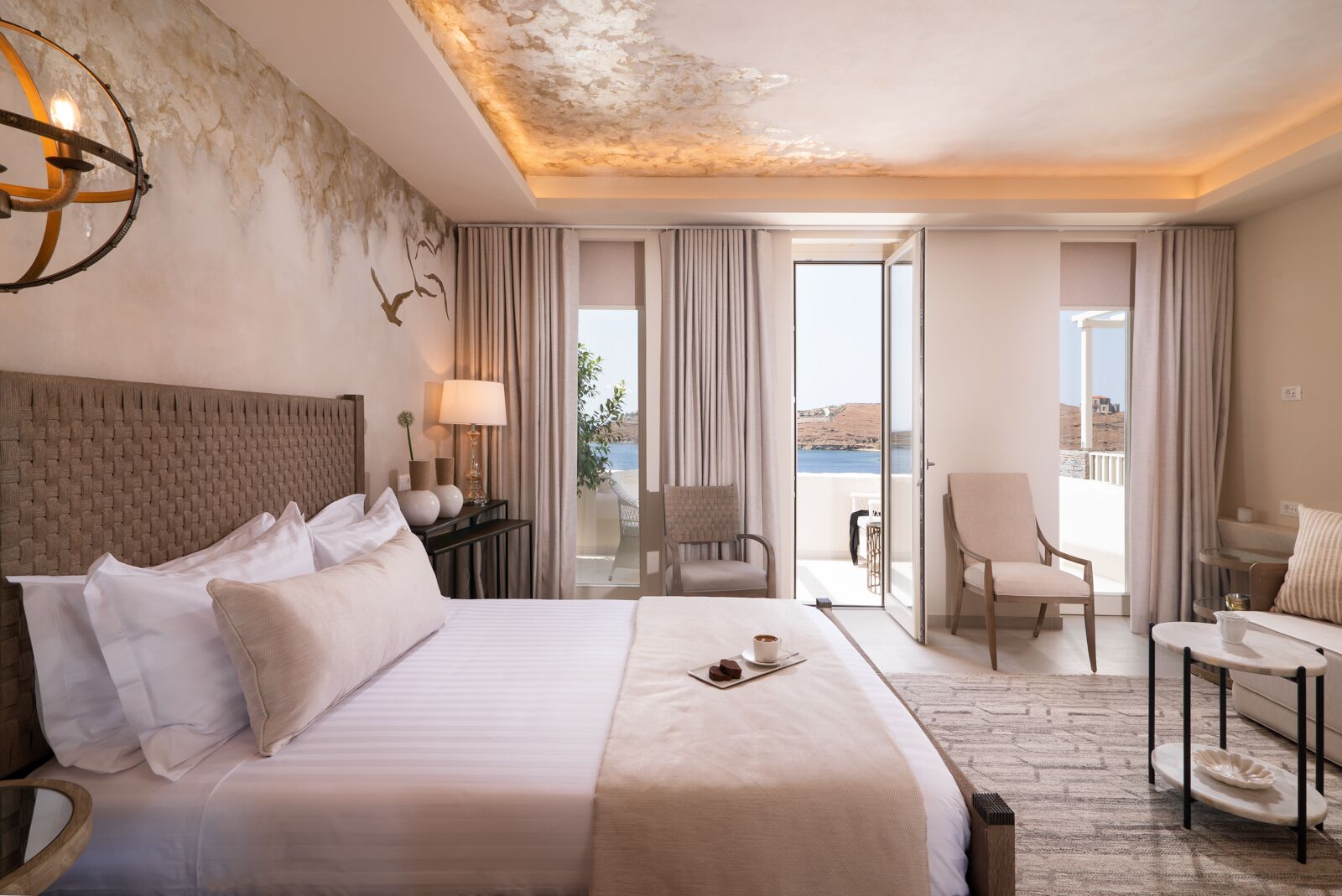 A hotel room with a large bed, a desk, a chair, and a balcony with a view of the sea.