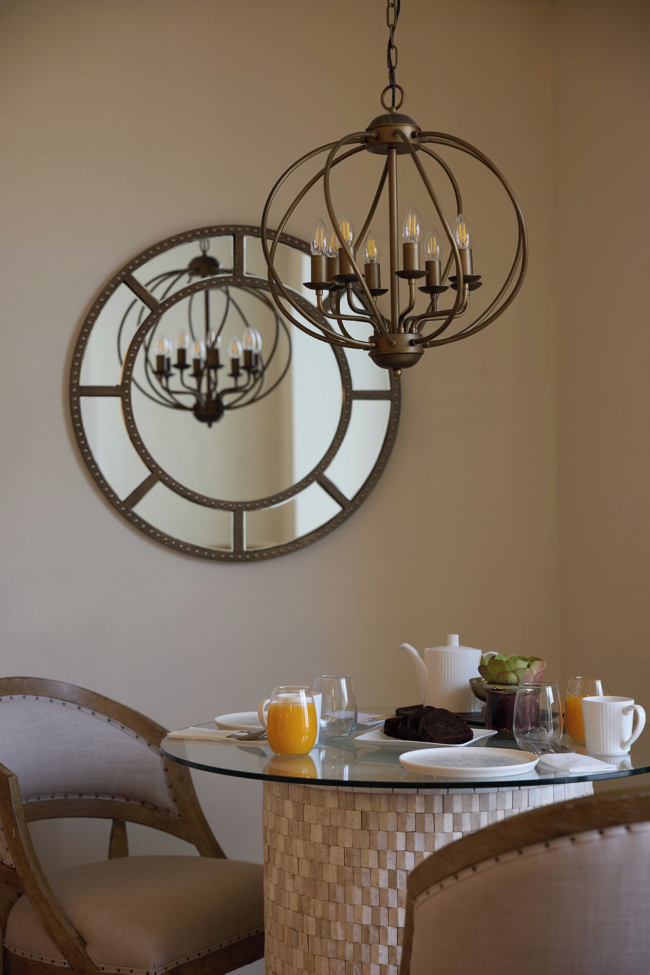 A dining area with a round table, chairs, a chandelier, and a large mirror on the wall.
