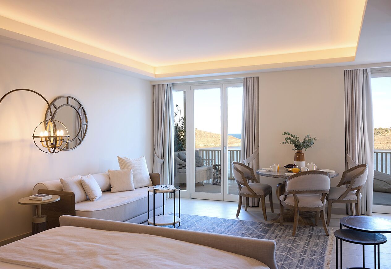 A hotel room with a bed, a sofa, a dining table with chairs, a small round table, and a balcony with a view.