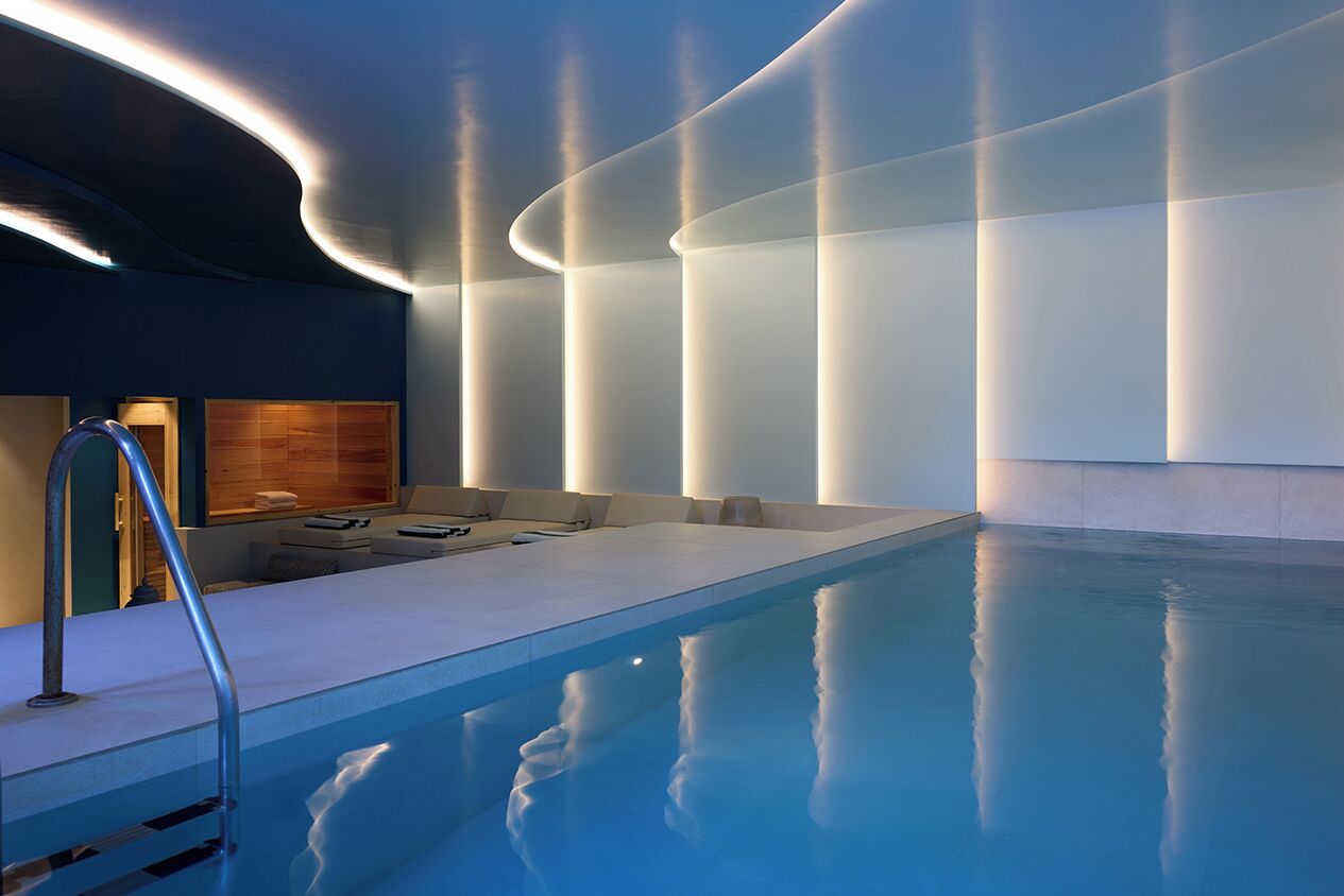 A modern indoor pool area with a clear pool, a seating area with cushions, and a wooden wall.