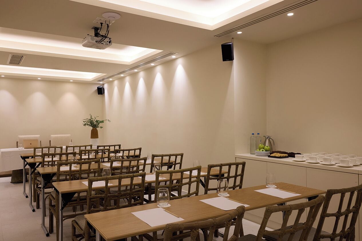 A conference room with multiple tables and chairs, a projector, a plant, and a small kitchenette area with cups and glasses.