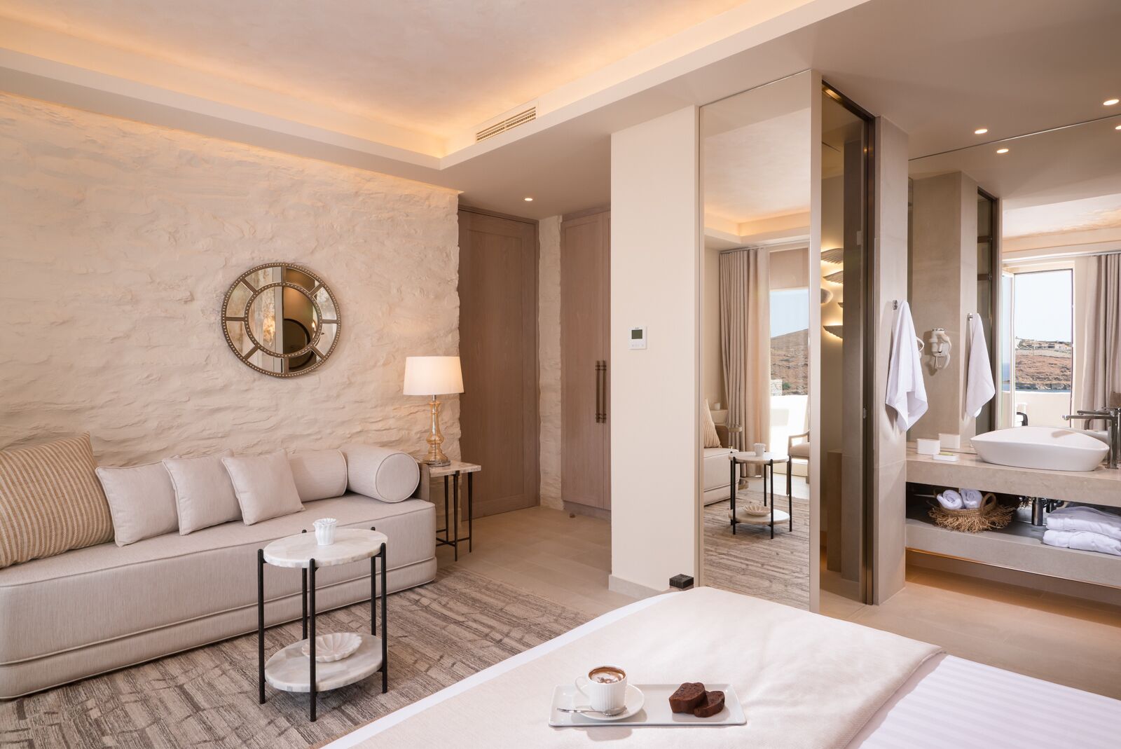 Una habitación de hotel moderna con una cama grande, un sofá, una pequeña mesa redonda con una taza y un plato, y un baño con un espejo grande y un lavamanos.