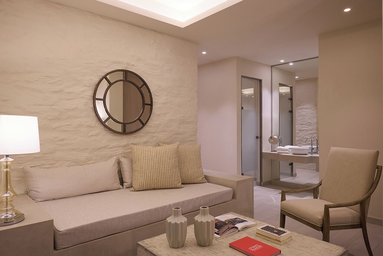 A modern living room with a beige sofa, a round mirror, a coffee table with books and vases, and a chair.