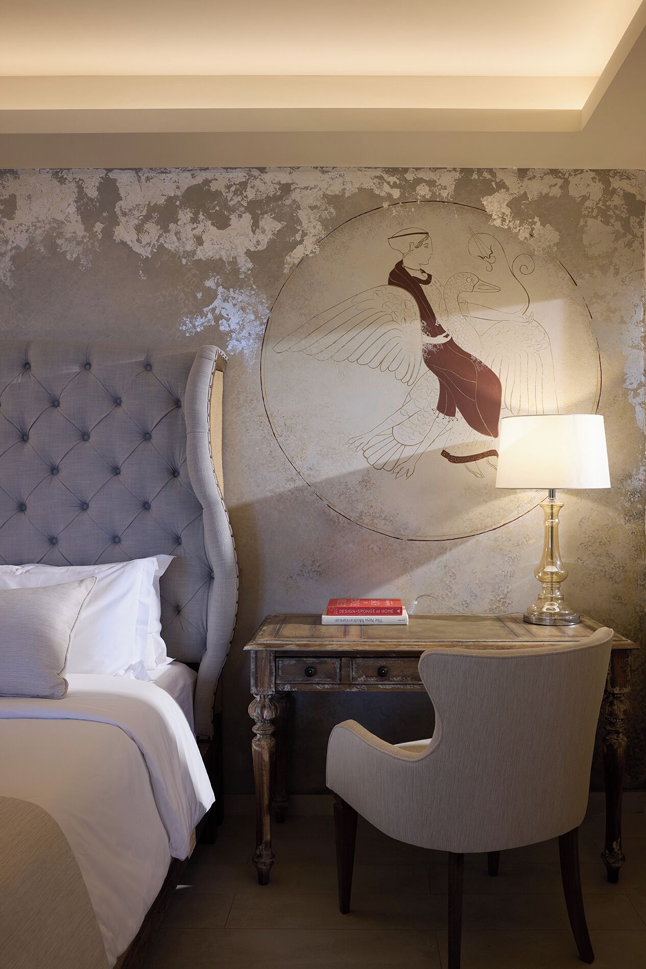 A hotel room with a tufted headboard, a bed with white linens, a desk with a lamp, and a wall mural.