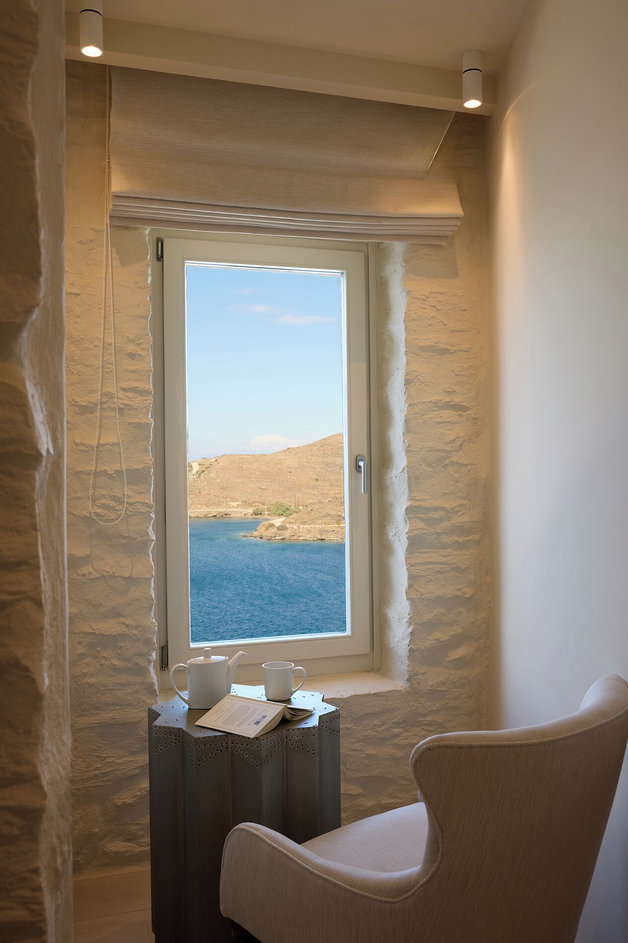A room with a view of the sea, a small table with a chair, and a window with a view of the landscape.