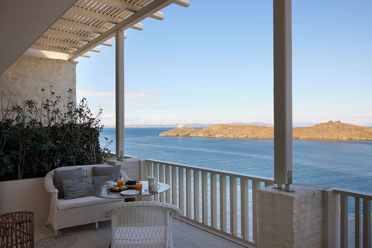 A balcony with a white wicker sofa, a small round table with a tray of food and drinks, and a view of the sea and distant hills.