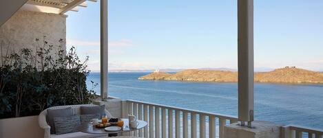 A balcony with a white wicker sofa, a small round table with a tray of food and drinks, and a view of the sea and distant hills.