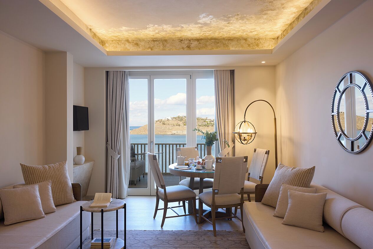 A modern hotel room with a balcony, a sofa, a dining table, and a large mirror.