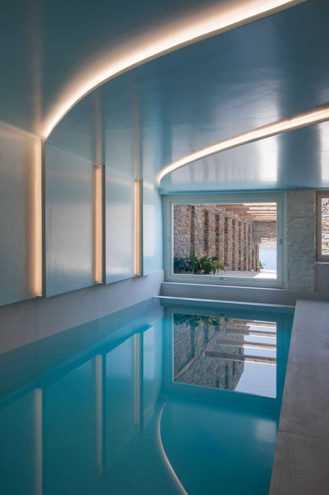 A modern indoor swimming pool with a curved ceiling and a view of an outdoor stone wall.
