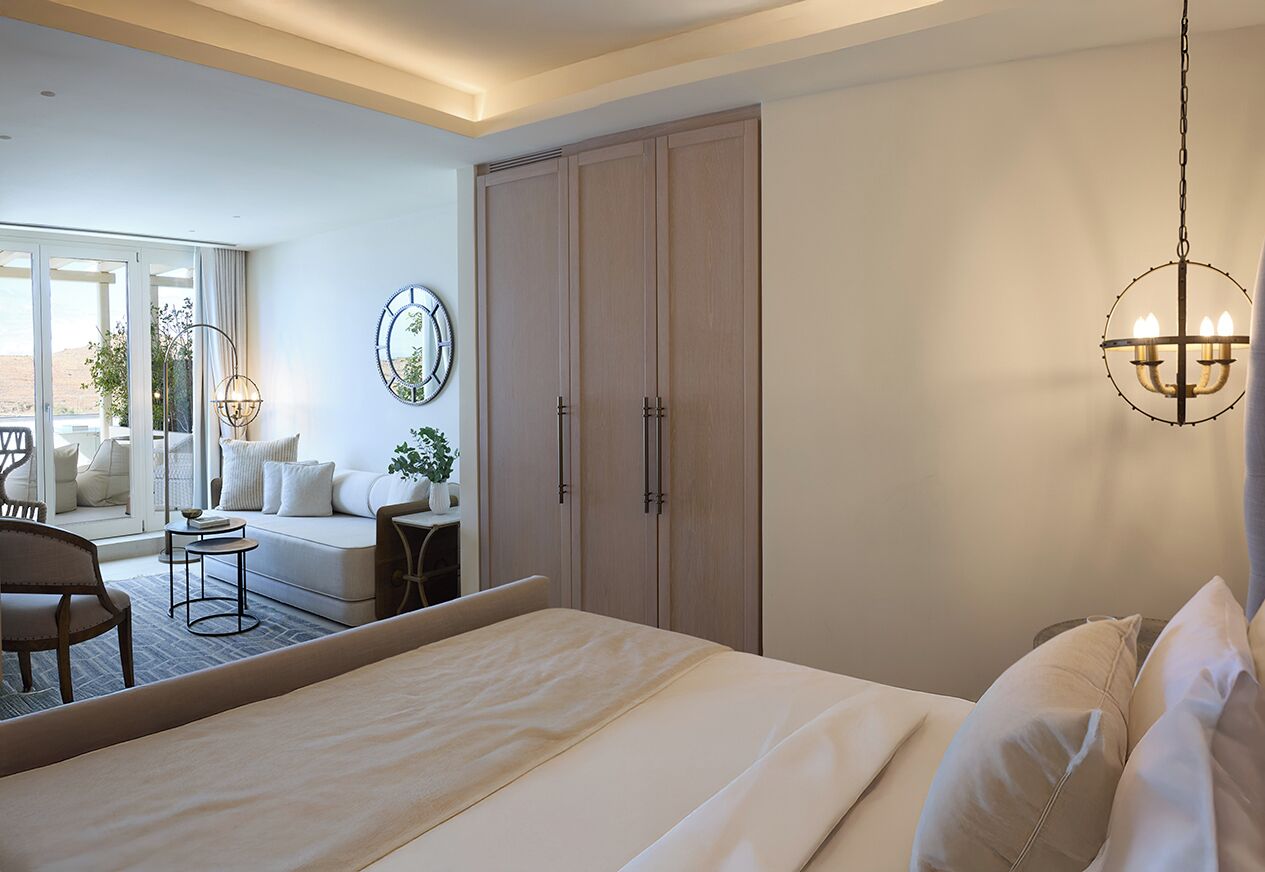 A bedroom with a large bed, a wardrobe, a chandelier, and a view of a balcony with outdoor furniture.