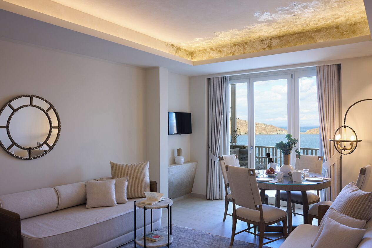 A modern living room with a sofa, a small round table, and a dining area with chairs. A flat-screen TV is mounted on the wall, and there is a large window with curtains.
