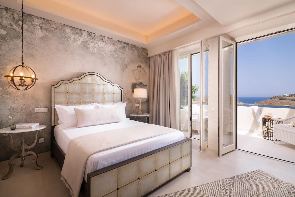 Een slaapkamer met een groot bed, een nachtkastje, een lamp en uitzicht op zee door een schuifdeur.