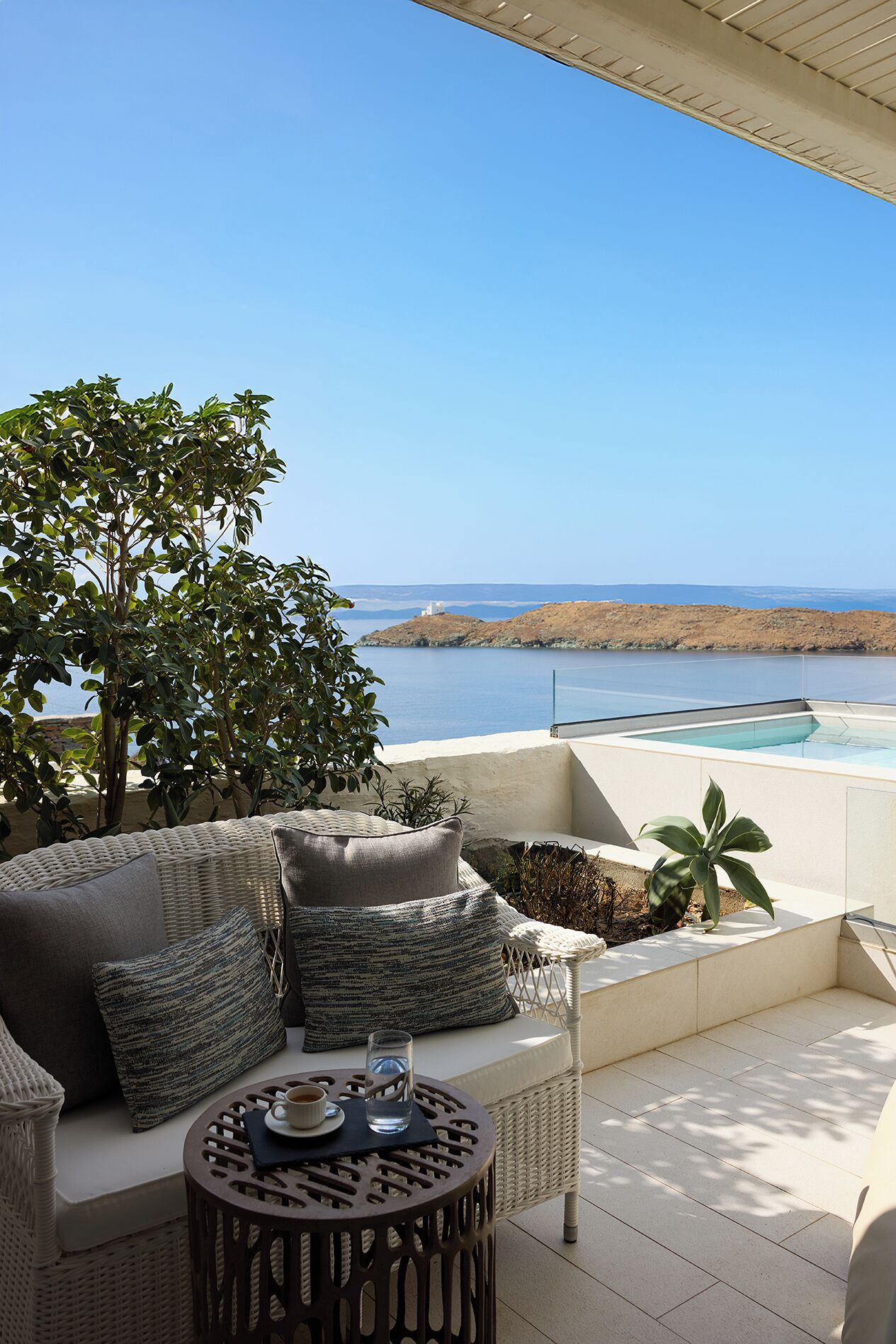 A terrace with a wicker sofa, a round table with a glass of water, and a view of a body of water and distant land.
