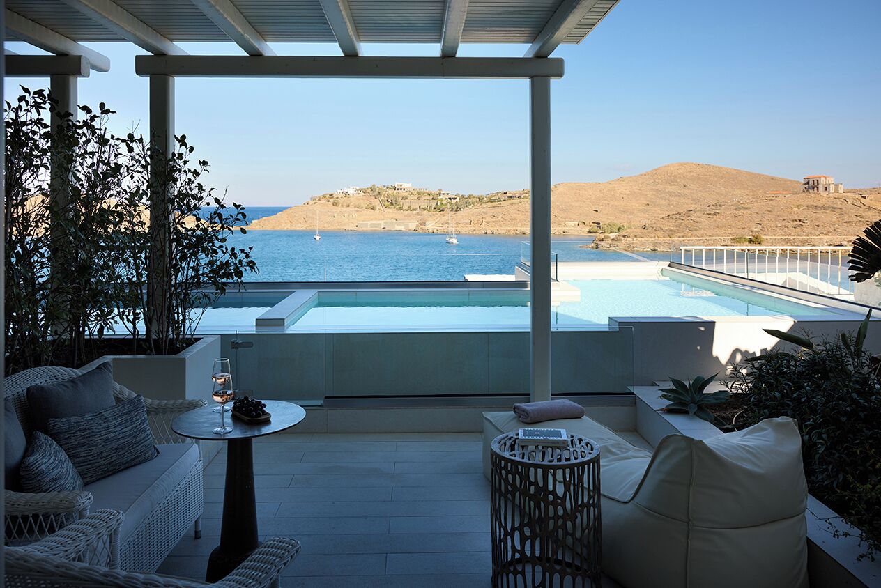 A terrace with a pool, wicker furniture, and a view of the sea and hills.