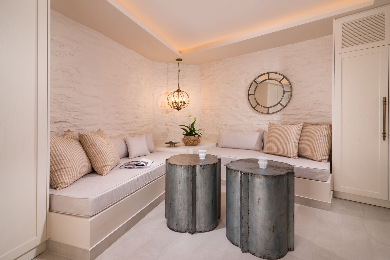 A modern living room with a white stone wall, a round mirror, a hanging light fixture, a sofa with cushions, and two round metal side tables.