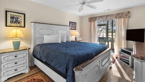 Une chambre à coucher comprenant un lit, deux tables de chevet, un ventilateur de plafond, une télévision et une fenêtre avec des rideaux.