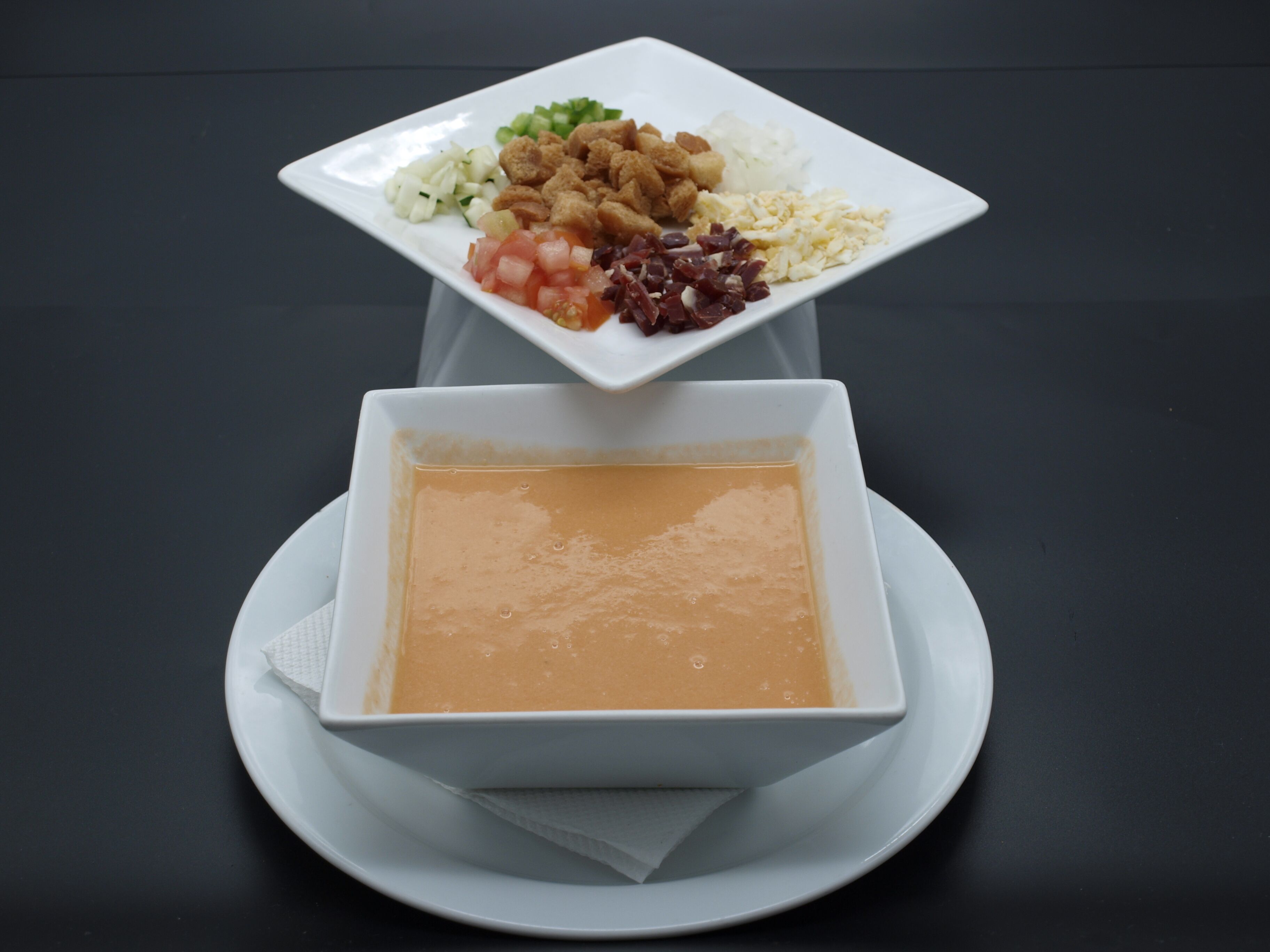 A white square plate with a variety of food items including diced tomatoes, onions, and a mix of meats, placed on a white plate with a bowl of soup underneath.