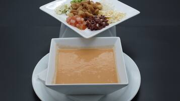 A white square plate with a variety of food items including diced tomatoes, onions, and a mix of meats, placed on a white plate with a bowl of soup underneath.