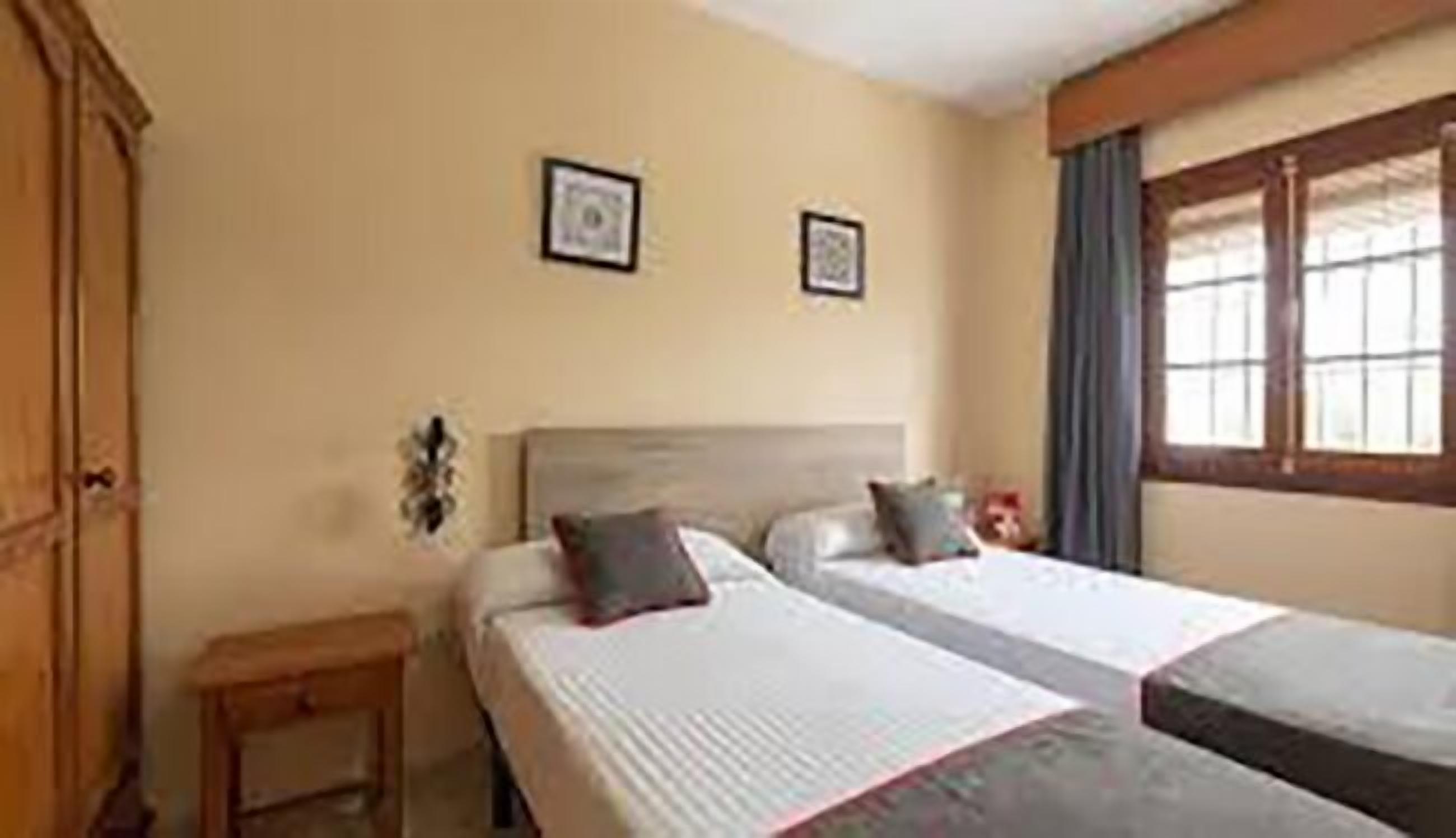 A hotel room with two beds, a wooden wardrobe, a small bedside table, and a window with curtains.