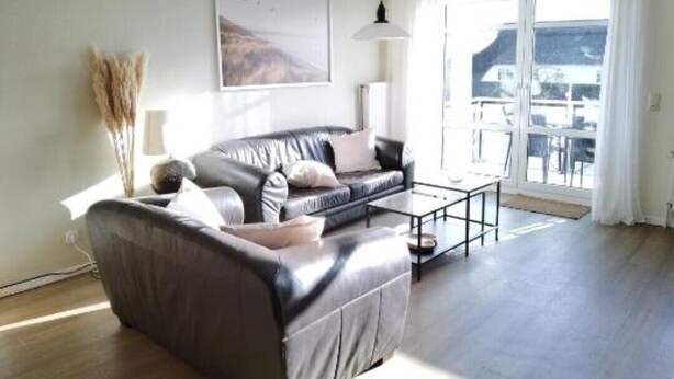 A spacious living room with a leather sofa, a coffee table, and a large window offering a view of the outdoors. - Schaake Hooksiel to feel good with 76sqm & balcony (Wangerland)
