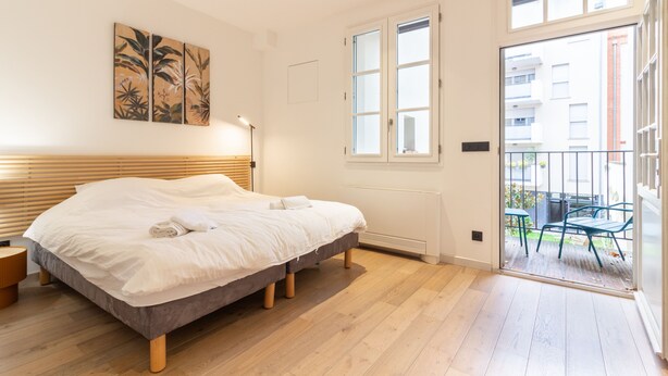 Ein Schlafzimmer mit einem Bett, einem Fenster und einer Tür, die zu einem Außenbereich mit Stühlen führt.