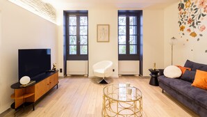 Ein modernes Wohnzimmer mit Flachbildfernseher, einem weißen Sessel, einem runden Couchtisch und einer Couch mit orangenen Kissen.
