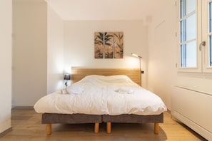 Uma cama bem arrumada com cabeceira de madeira, uma luminária na parede e uma obra de arte com tema tropical acima.
