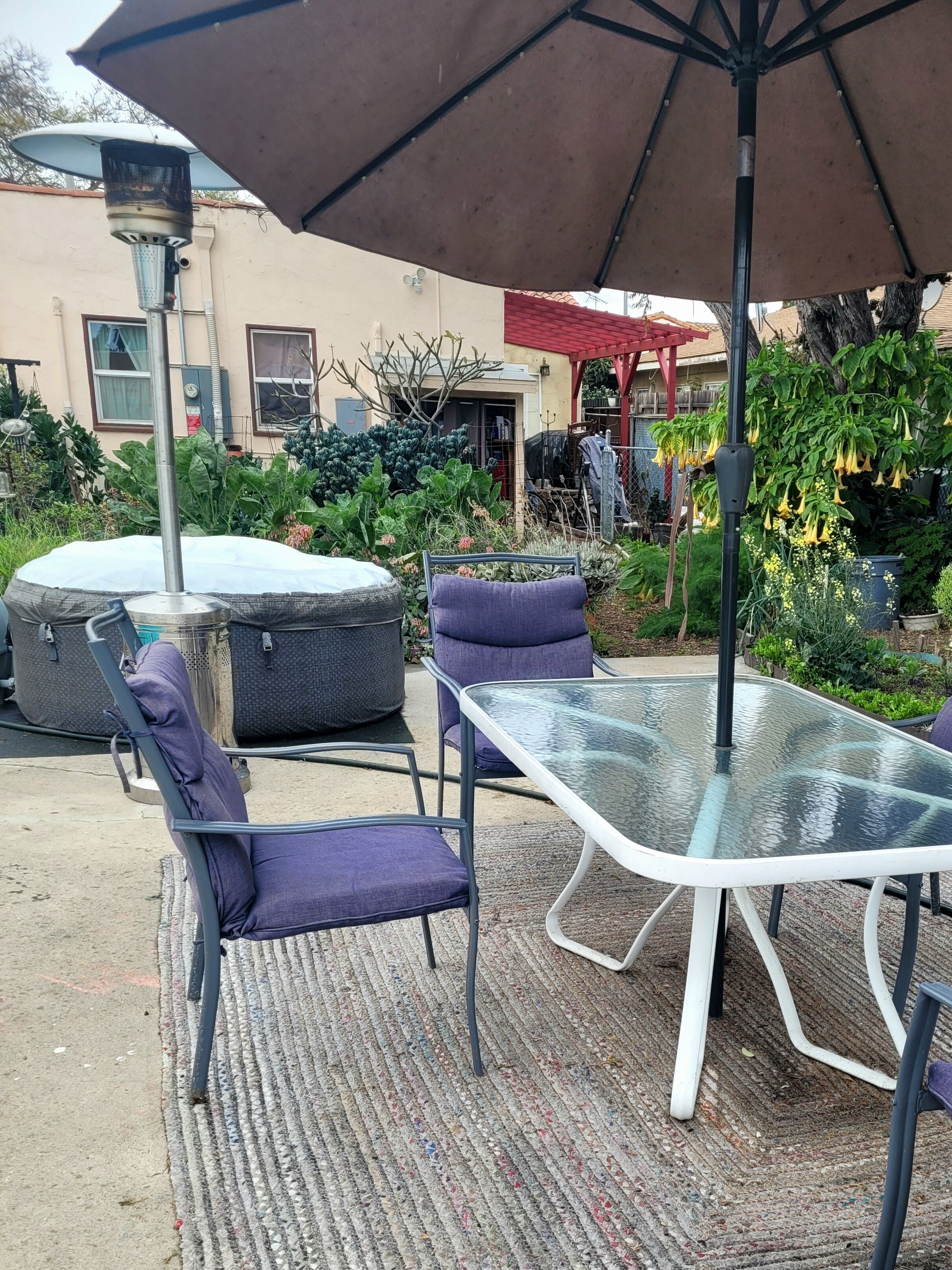 Eine Terrasse im Freien mit einem Glastisch und violetten Stühlen, umgeben von Grünpflanzen und einem Gebäude im Hintergrund.