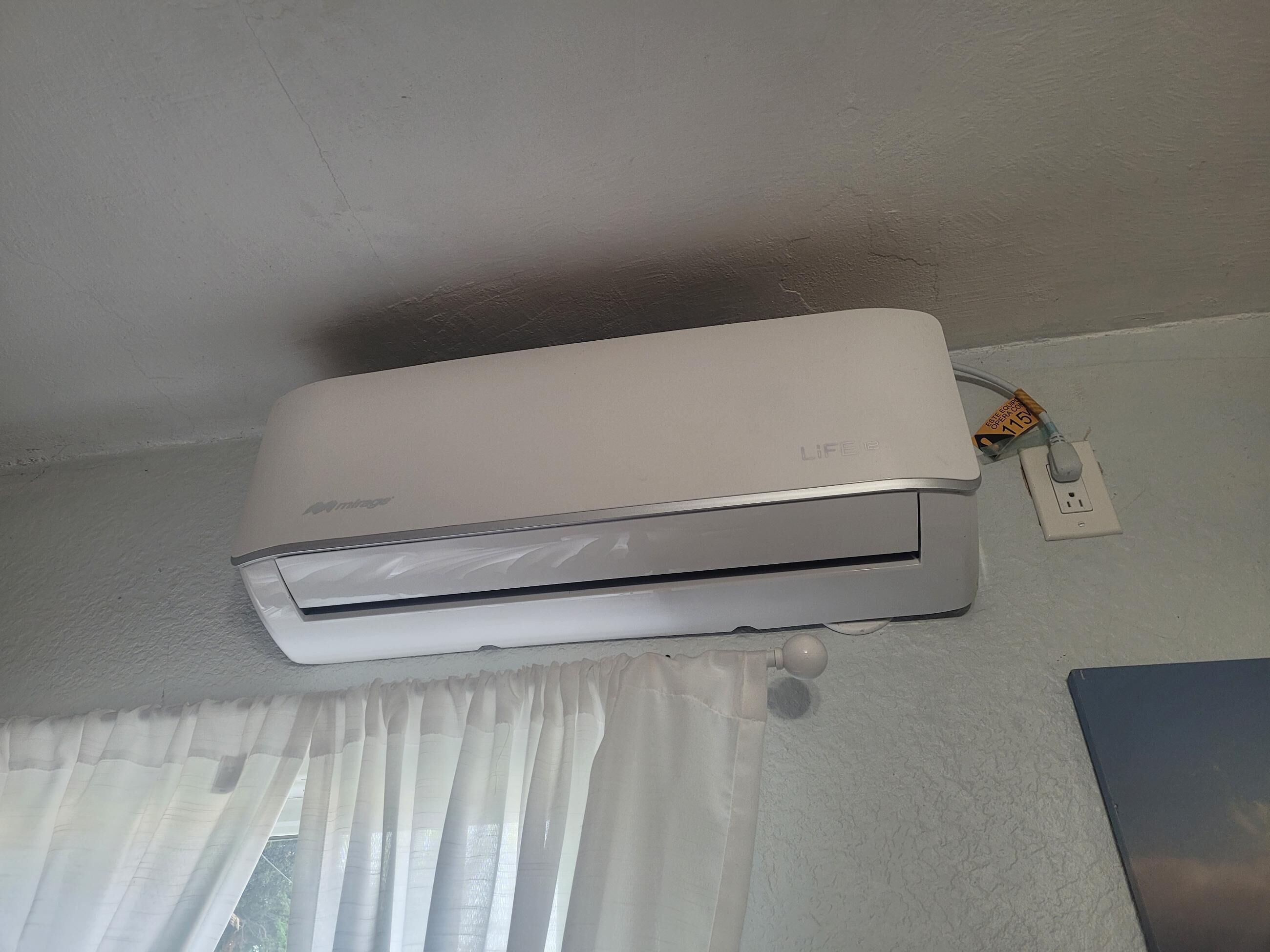 A white air conditioner mounted on a wall with a power cord plugged into a wall socket.