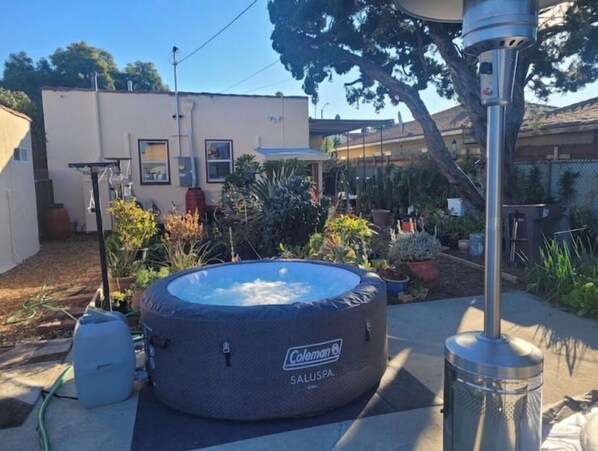 A Coleman SALUSPA hot tub in a backyard with a house, potted plants, and a metal heater.