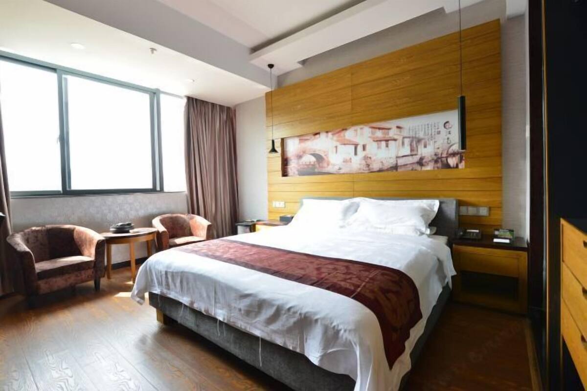 Hotellihuone, jossa on suuri sänky, puinen pääty, pieni pöytä, kaksi nojatuolia ja suuri ikkuna, jossa on verhot.