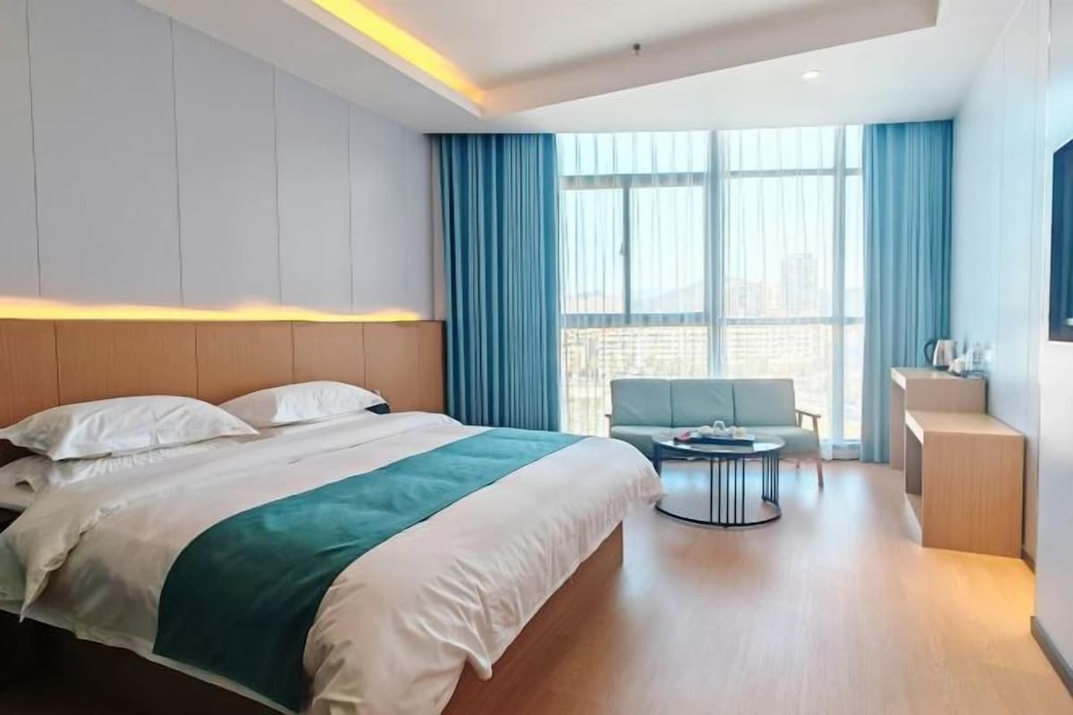 Habitación de hotel moderna con una cama grande, un área de descanso con una mesa y vistas de la ciudad a través de grandes ventanales.
