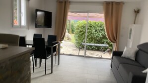 Un salon moderne comprenant un canapé, un téléviseur fixé au mur, un coin repas avec des chaises et une table, ainsi qu’une porte-fenêtre coulissante donnant sur une terrasse avec une table et des chaises.