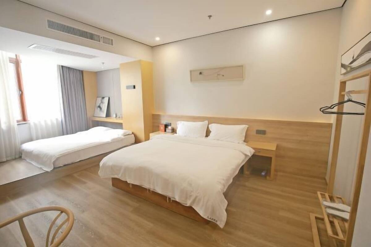 Una habitación de hotel con una cama grande, una cama más pequeña, piso de madera, una mesita de noche y un estante en la pared.