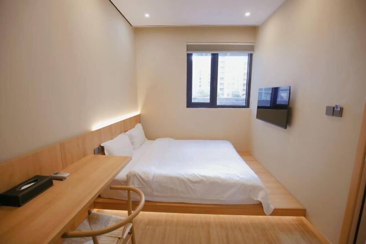 Habitación de hotel compacta con una cama individual, una mesita de noche, una silla, un televisor montado en la pared y una ventana que ofrece luz natural.
