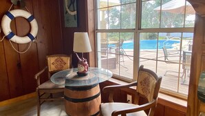 Une pièce avec une table en verre, des chaises en bois, une lampe, un gilet de sauvetage et une vue sur une piscine et du mobilier d’extérieur.