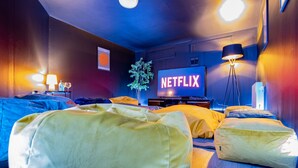 Une chambre d’hôtel dotée d’un grand téléviseur à écran plat affichant le logo « NETFLIX », d’un lit avec des oreillers jaunes, d’une table de chevet avec une lampe et d’une plante.
